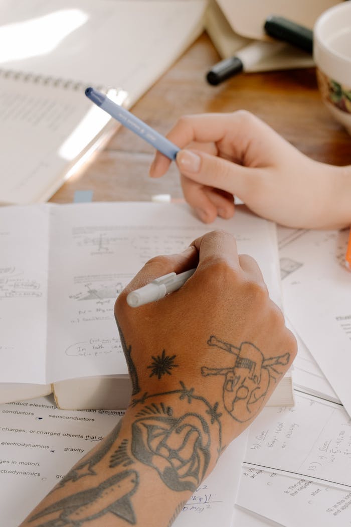 Close-up view of tattooed and non-tattooed hands holding pens while studying from textbooks.