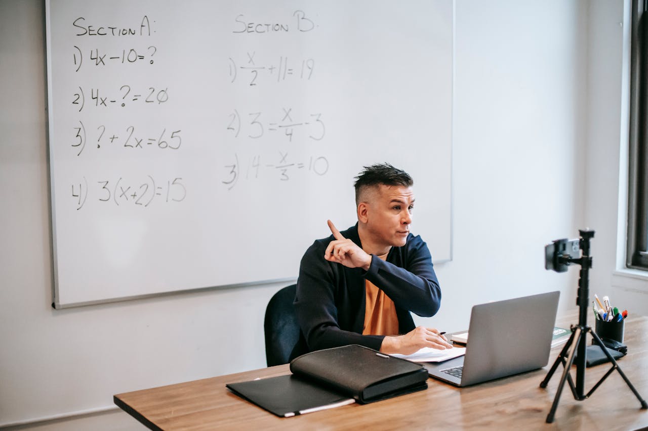 A male teacher explains math equations during an online class, using a laptop and whiteboard.